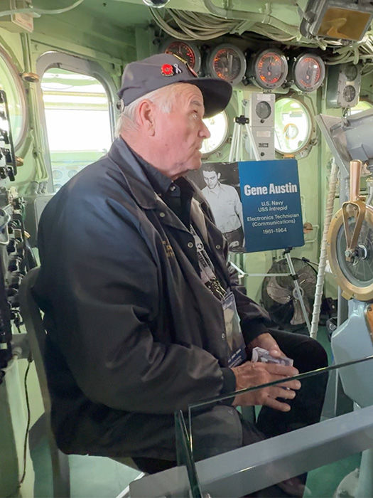 Elderly veteran in a dark jacket and cap sits inside a submarine-like control room beside a Gene Austin Navy exhibit sign.