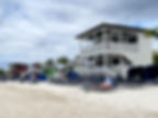 Beach scene with colorful cabanas, blue lounge chairs, and a two-story white house. Cloudy sky and palm trees in the background.
