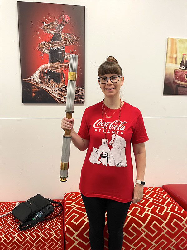 A woman in a red "Coca-Cola Atlanta" shirt holds an olympic torch. Background features Coca-Cola art with red and black tones.