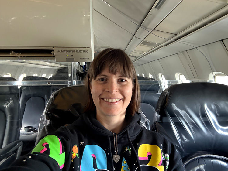 Smiling woman takes a selfie in an airplane cabin, wearing a black hoodie with colorful 2023 print; Mitsubishi Electric unit overhead.