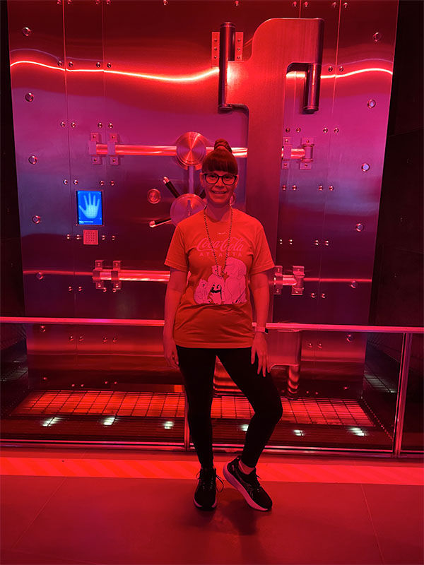 Woman in a red coca cola shirt stands in front of a large, illuminated vault door with red lighting. A blue handprint scanner is visible.