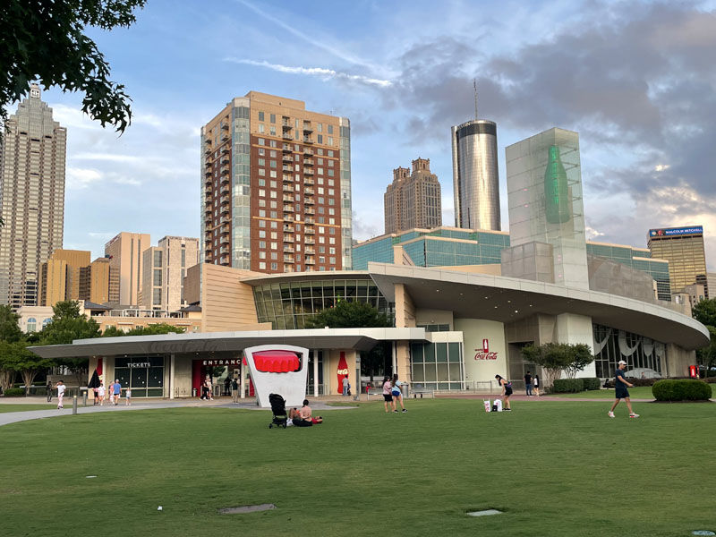 People relax on a green lawn in front of modern buildings and the Coca-Cola museum. A large red bottle cap and a glass bottle icon are visible.