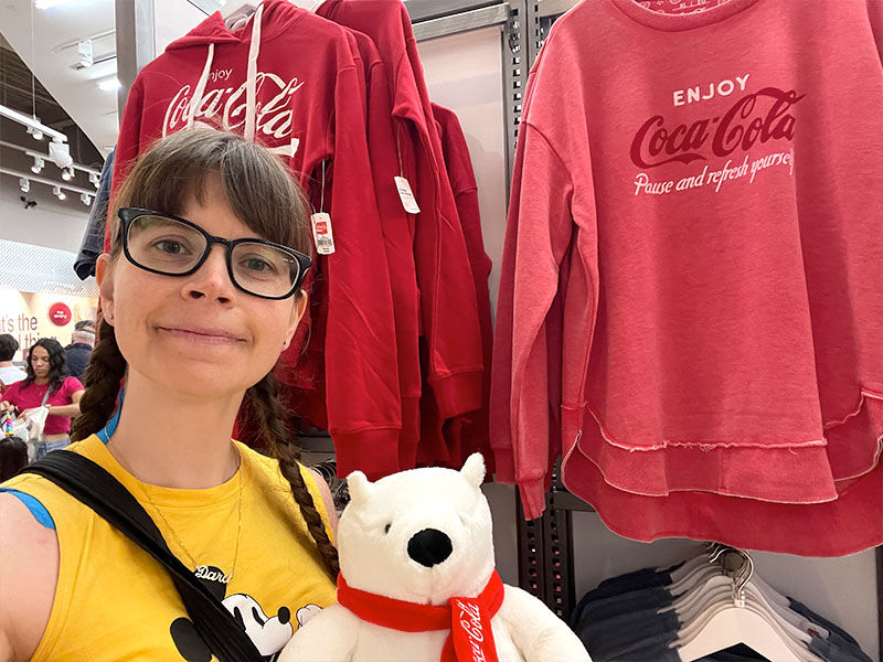 Woman in yellow Mickey Mouse shirt holds a white stuffed polar bear. Red Coca-Cola hoodies in background. Store setting, relaxed mood.