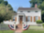 White colonial house with brown shutters, a picket fence, and a patriotic bunting. Green lawn and trees surround the sunny setting.