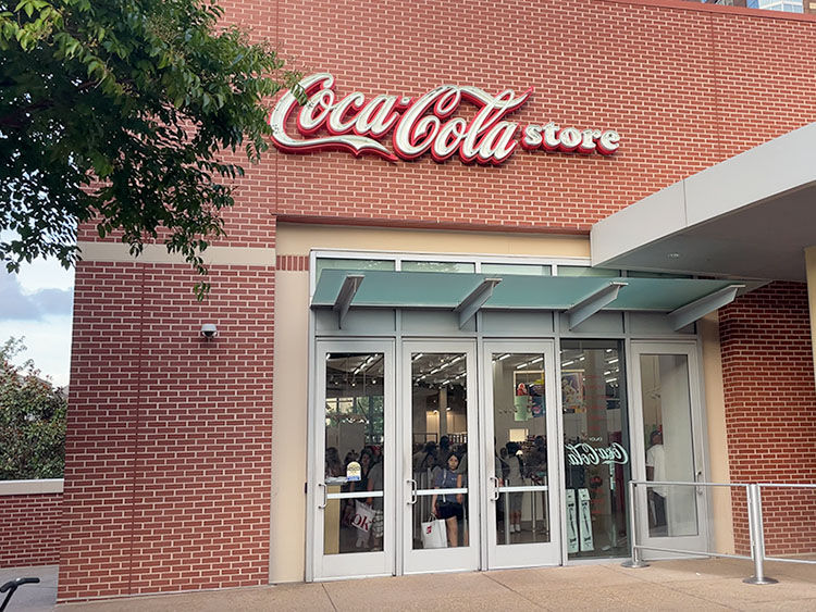Coca-Cola store entrance with glass doors on a red brick building. Green awning above, and visible logo. People inside. Bright mood.