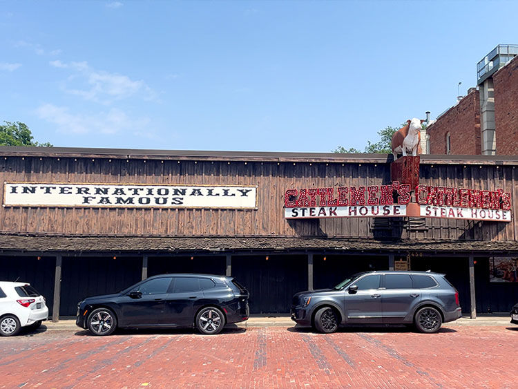 Street view of a steakhouse with wood exterior, signs reading "INTERNATIONALLY FAMOUS" and "STEAK HOUSE." Parked cars in front, sunny day.