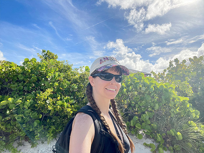 Woman with braided hair, cap, and sunglasses smiles on a sunny beach path. Green foliage and blue sky with clouds in the background.