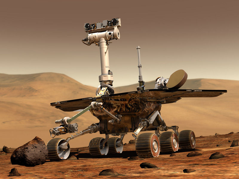 Mars rover on a rocky red desert, showing its camera mast and solar panels under a hazy orange sky.
