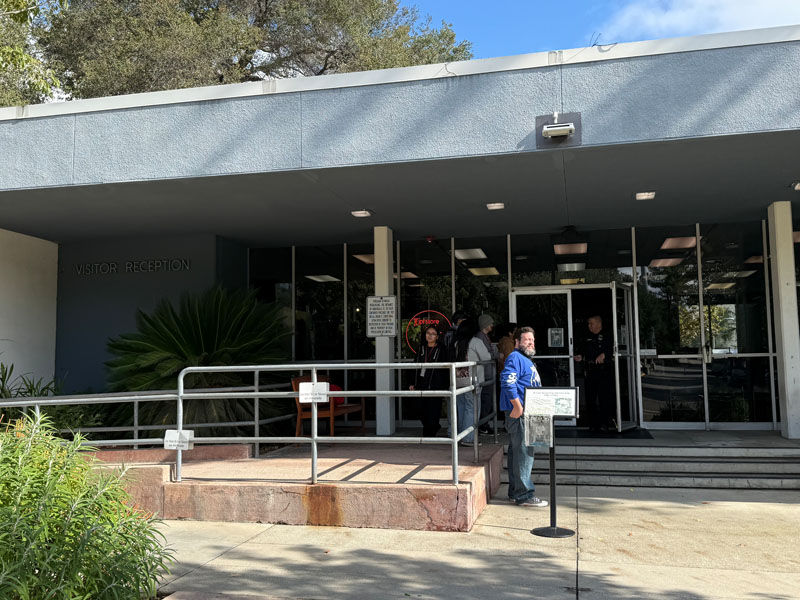 People stand outside a VISITOR RECEPTION entrance with glass doors; a man in a blue shirt waits by a sign on a sunny day.