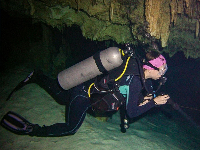 Scuba diver in a cave swims low over the rocky floor, wearing a tank, mask and pink headband, with a flashlight in dark water.