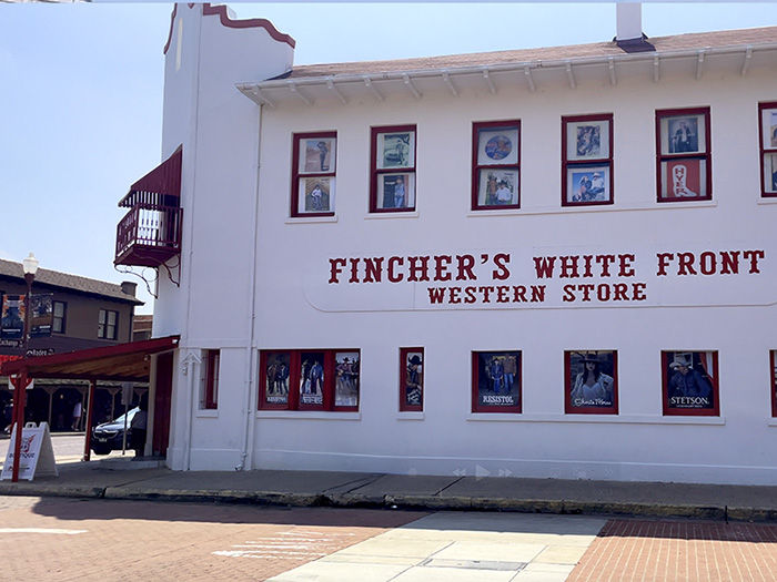 White building with "Fincher's White Front Western Store" in red letters, displaying western wear posters in windows, set on a sunny street.