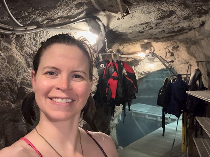 Smiling person in a cave-like setting with dim lighting. Life jackets hang on the rocky walls; shelves line the tunnel. Mood is adventurous.