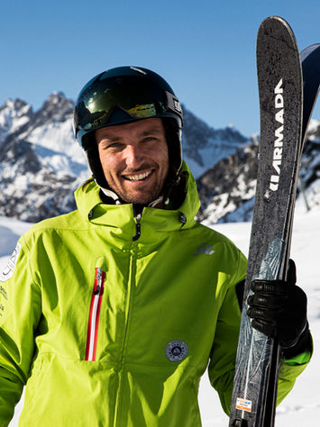 Skier smiling in the mountains, green jacket, helmet, Armada skis, winter landscape.