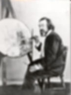 Painter Richard Dadd, seated, focusing intently, adds detail to a vibrant, circular artwork on an easel. Monochrome setting with a reflective mood.