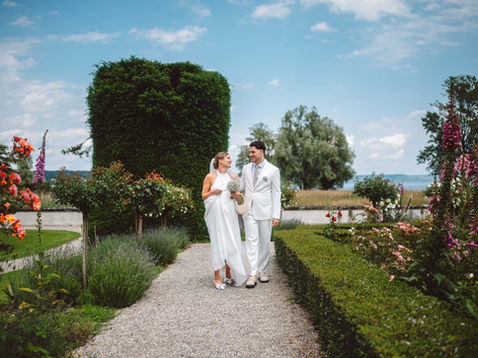 Braut und Bräutigam in weißen Outfits spazieren Hand in Hand durch den blühenden Garten der Seeburg Kreuzlingen mit Blick auf den Bodensee, umgeben von farbenfrohen Blumen und sommerlichem Licht.