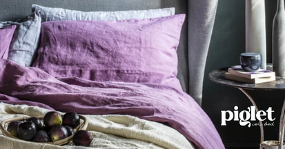 Purple bedding with pillows, a basket of figs on a cream blanket, and a side table with books and a cup. Piglet in Bed.