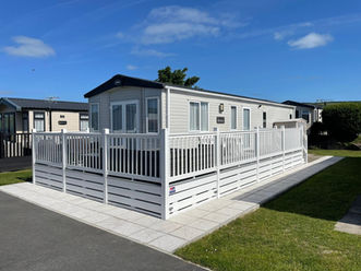 A white caravan with a black roof and a white plastic decking. The caravan has a small path on the front and side. Clear blue sky in the background.