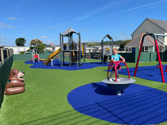 Children's outdoor play park with green and blue padded flooring. There is a young girl having fun and playing on a small round piece of park equipment. 
