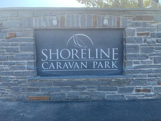 Grey Brick Wall with a Black Slate Sign that reads "Shoreline Caravan Park" with a small illustration of a Wave both in White.