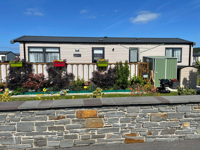 A white caravan with a black roof and a dark wood decking. The decking is decorated with shrubs and potted plants attached. There is a grey and brown brick wall in the foreground.