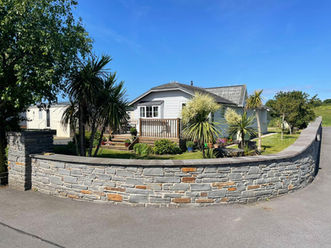 Small White Bungalow with a grass front garden and palm trees outside. There is a grey and orange brick wall in front of the property.
