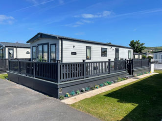 A white caravan with a black roof and a black decking. The caravan has a small path on the side with some flowers planted along the path. Clear blue sky in the background.