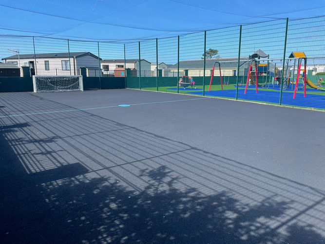Netted football court with a grey floor and a white metal goal. The court is surrounded by green metal railings.