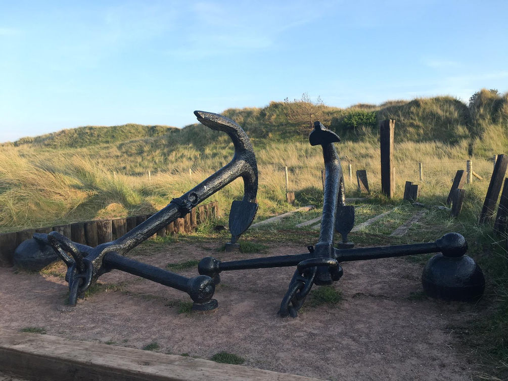 Two Large Black Metal Anchors side by side with green and beige sand dunes in the background.
