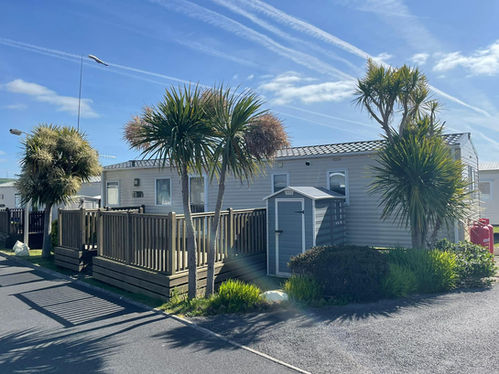 A white caravan with a natural wood decking and a small grey shed on the front. There are small palm trees around the caravan. Cloudy blue sky in the background.