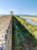 A picture of a beach. A grey brick wall along the left side of the photograph. There is a white lighthouse with a red roof at the end of the wall. A grass path leading up to the lighthouse. Blue sea and sky in the background.