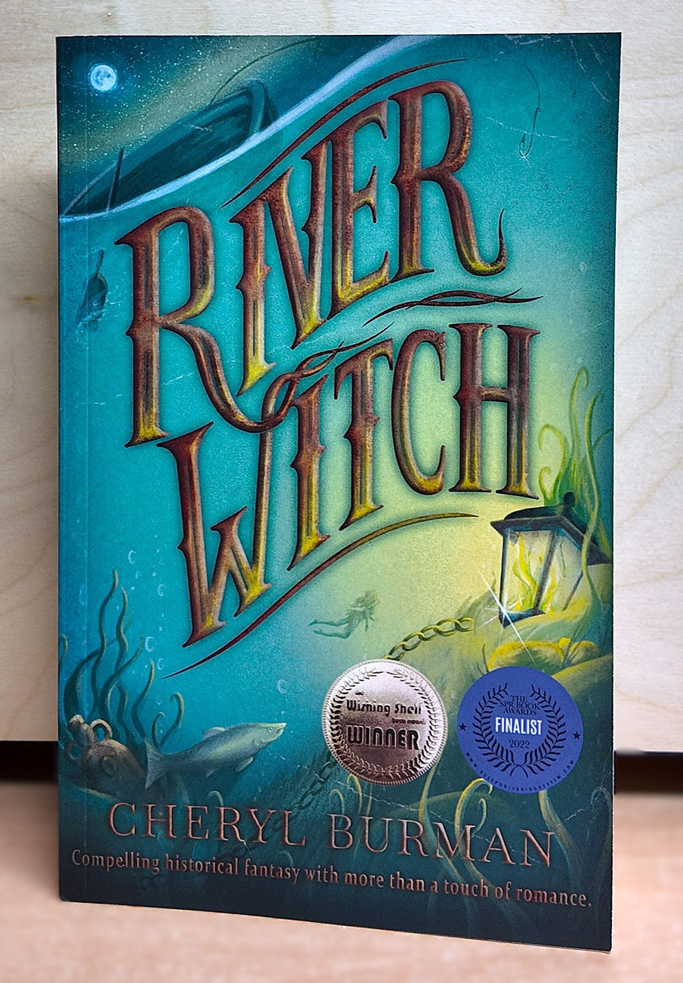 Book "River Witch" by Cheryl Burman. Blue cover with fish, lantern, and moon. Silver "Winner" and blue "Finalist" stickers displayed.