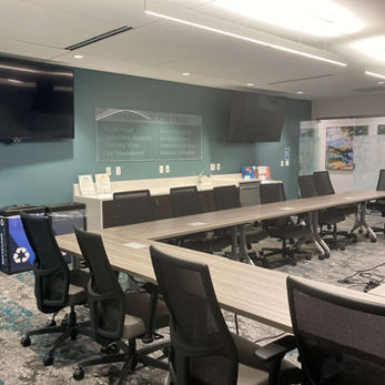 A large room with tables in the shape of an oval. About 3/4 of the oval is visible and at an angle. On the outside of the oval are high backed chairs with wheels and armrests. There are 2 TV monitors on the left side of the room, in addition to a long counter that is between the monitors. To the left of the counter are trash and recycling.