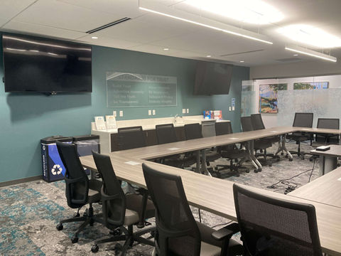 A large room with tables in the shape of an oval. About 3/4 of the oval is visible and at an angle. On the outside of the oval are high backed chairs with wheels and armrests. There are 2 TV monitors on the left side of the room, in addition to a long counter that is between the monitors. To the left of the counter are trash and recycling.