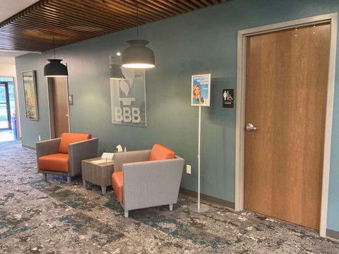 An open area with two orange armchairs flanking a table. There are hanging lights over the chairs. On either side of the chairs is a door for the restroom.