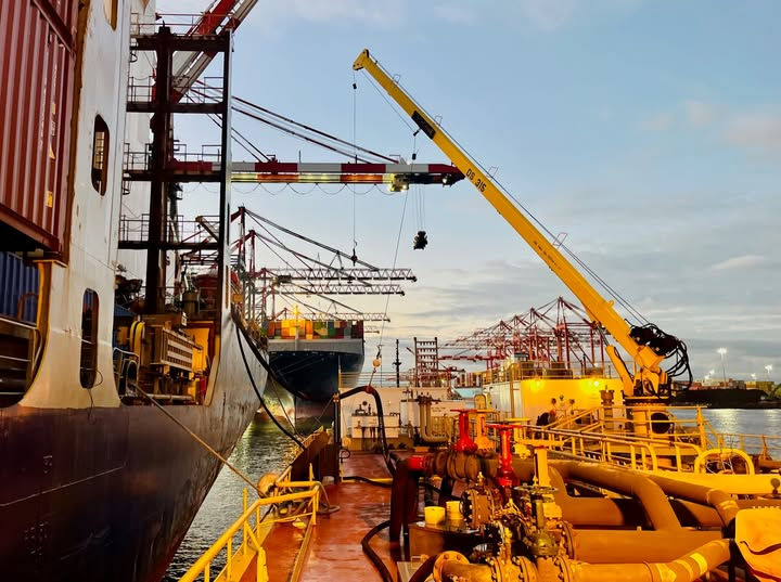 Cargo ships bunkering at sunset. Cranes and hoses are visible, creating a vibrant industrial scene under a colorful sky.
