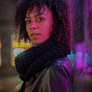 Das Porträt zeigt eine junge Frau in einem schulterfreien, bodenlangen, schwarzen Abendkleid mit einer taillierten Silhouette. Ihr glattes, dunkles Haar fällt in sanften Wellen über ihre Schultern. Sie hat dezentes Make-up mit betonten Augen und roten Lippen. Ihr Blick ist selbstbewusst und leicht nach links gerichtet. Das Umgebungslicht betont die eleganten Konturen ihres Gesichts und hebt die feinen Details ihres Kleides hervor, einschließlich der feinen Stickereien und des asymmetrischen Ausschnitts. Im Hintergrund ist eine neutrale, leicht texturierte Wand zu erkennen, die das Hauptaugenmerk auf das Model und ihr stilvolles Outfit lenkt