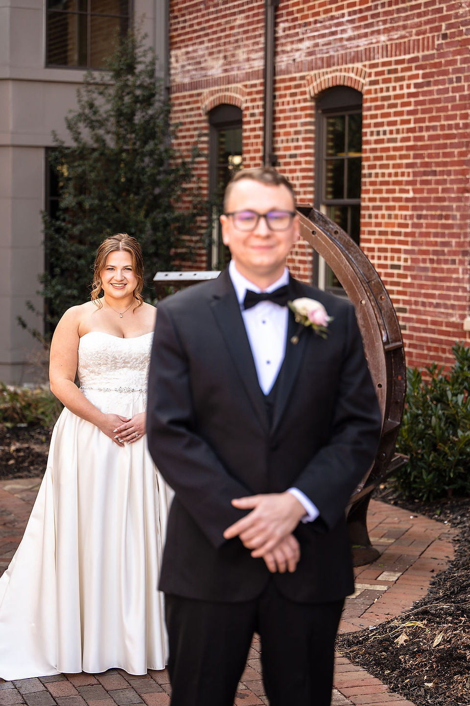 a groom in a black suit smiling at the camera with a bride  in the back in a wedding gown smiling at the camera at the Wilbur Hotel in Lititz Pennsylvania
