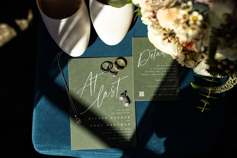 flat lay of wedding invitation, wedding rings, earrings, shoes, and a bouquet against a blue velvet 