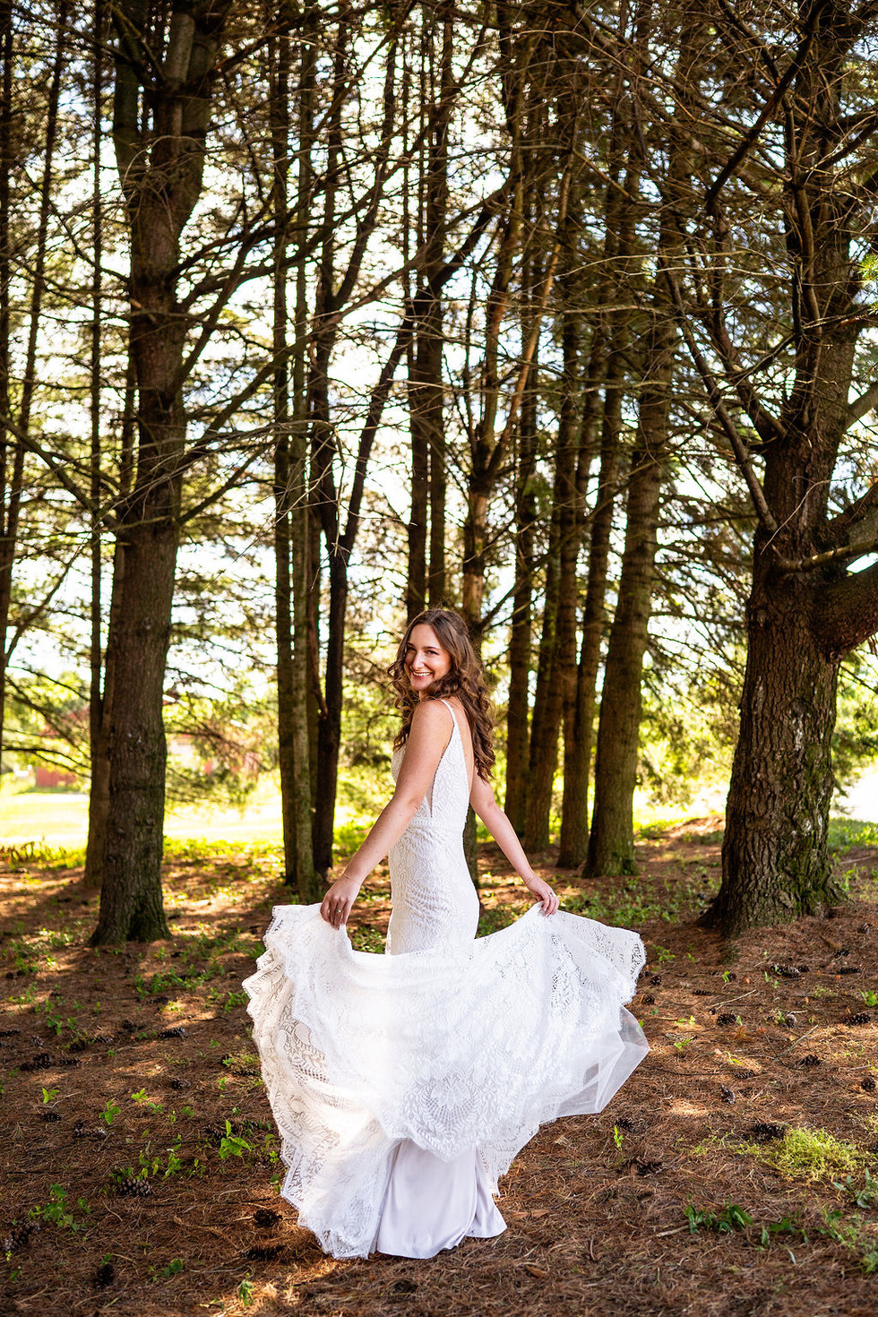 a woman with brown hair smiling and twirling in a white lace bridal gown surrounded by tall trees at Reinhart's Barn on Oak Lane in Bernville, Pennsylvania