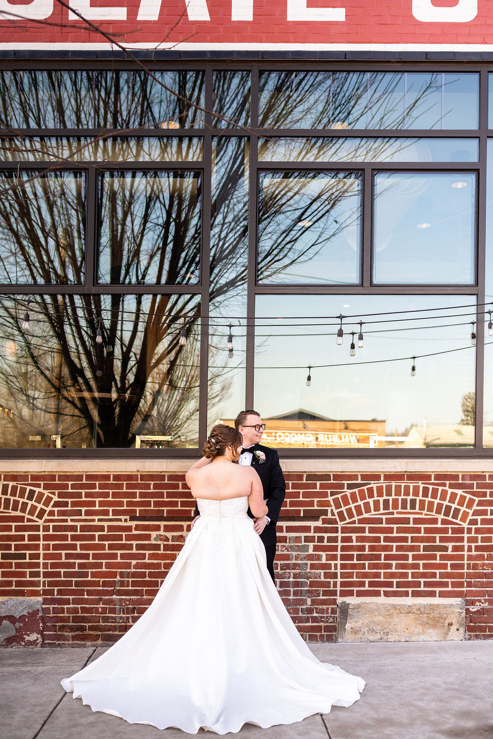 view of the back of a wedding dress and a groom posing in front of a brick building with windows  in Lititz Pennsylvania