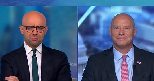 Split-screen TV interview on Bloomberg showing two men in dark suits: on the left, a bald man with dark-rimmed glasses and a green tie in the studio; on the right, a bald man with a pink tie and an American flag lapel pin, smiling, appearing remotely with a Washington, D.C. backdrop.