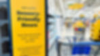 Sign for "Sensory-Friendly Hours" in a store, yellow with black text. Shopping carts and shelves visible in bright aisle background.