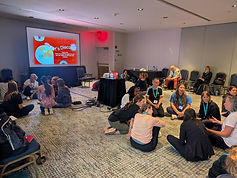 Large presentation room with a projected image on the screen and adults sitting in on the floor in groups