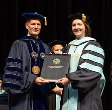 Man and woman in dark blue doctoral graduation garb holding a dark blue diploma holder smiling at camera.