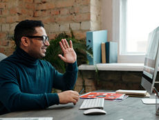 Virtual Assistant with glasses waves during a work call on a computer in a cozy home office.