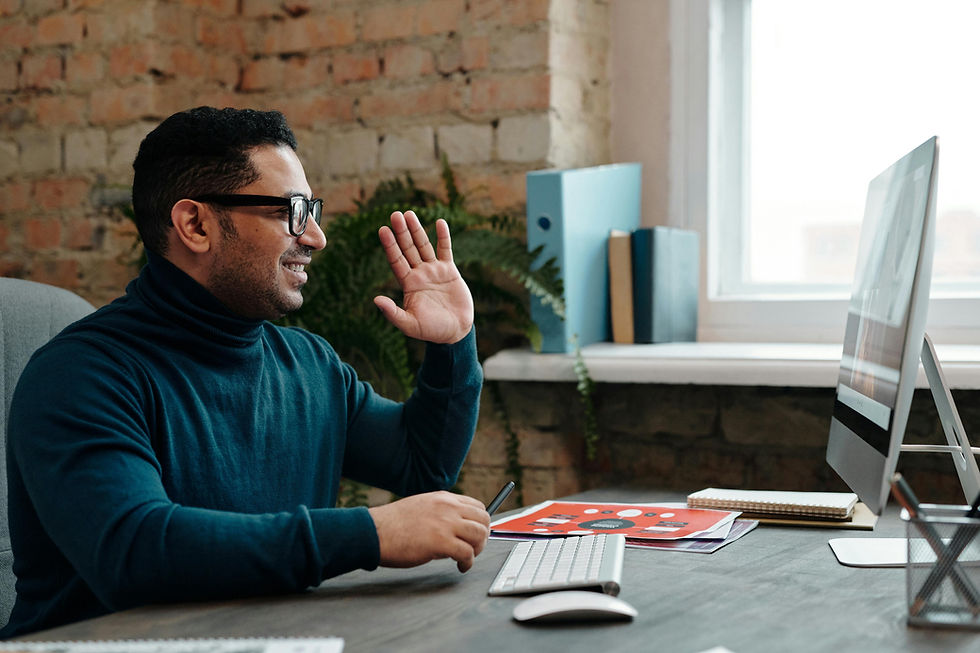 Virtual Assistant with glasses waves during a work call on a computer in a cozy home office.