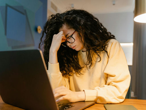 A tired Virtual Assistant with curly hair and glasses experiencing stress while working on a laptop.