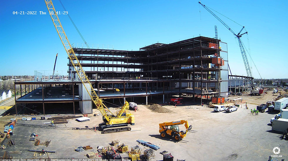Construction site of future Lutheran Medical Center, white trucks, storage units, construction materials staged out on the ground, two cranes, one yellow one red, yellow fork lift, dumpsters, and construction workers in PPE