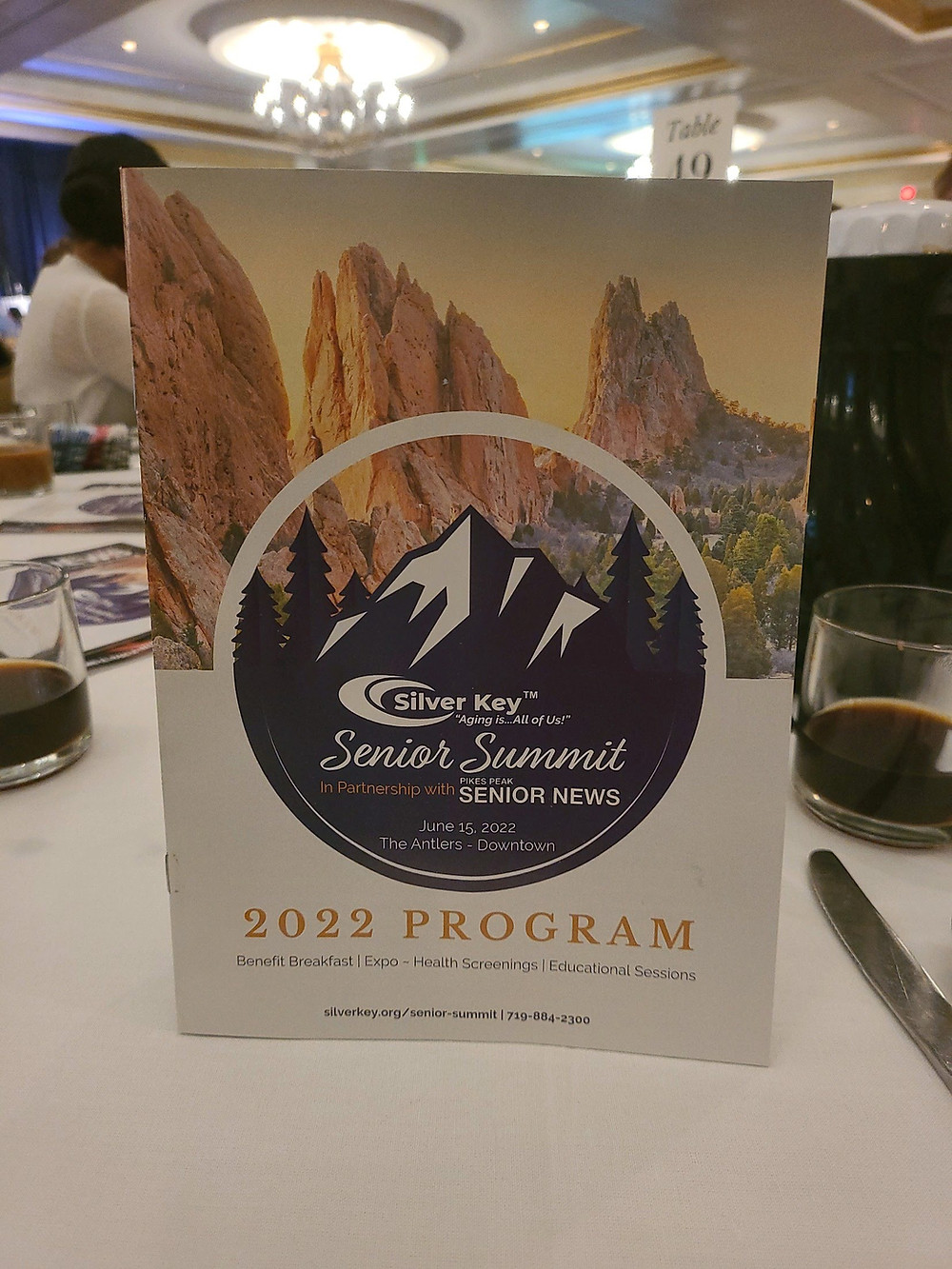 Senior Summit Program cover page on table at the event, white table cloth and sign Table 19, clear coffee mug with coffee, background conference room with white ceiling with gold accents and lighted chandelier