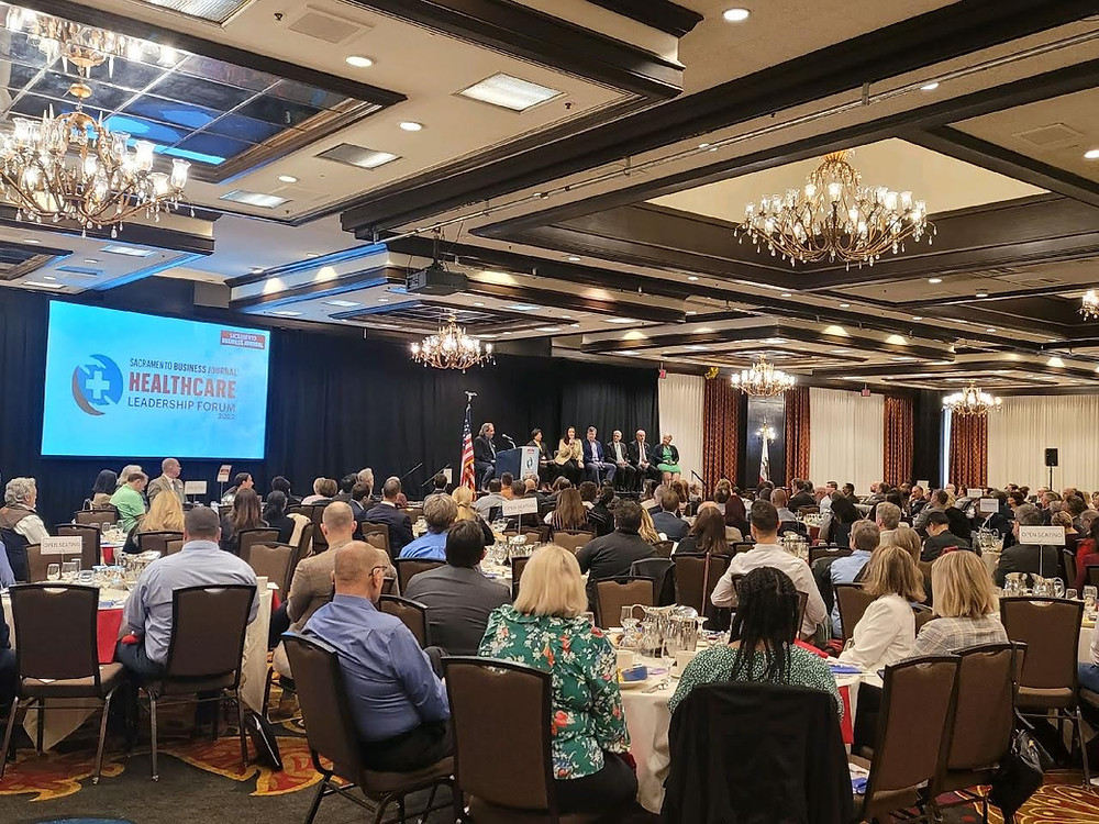 Hotel conference room, panelists on stage with screen displaying Sacramento Business Journal Healthcare Leadership Forum 2022 - attendees sitting at round tables focused on panelists and moderator- panelists are seated with wireless microphone - moderator is seated at podium with microphone- American flag is also on the stage - background includes crystal, glass, and gold chandeliers hanging from white and brown ceiling, walls with red, black, and white drapes, carpeted multi-colored flooring, people in business casual attire, round tables with white linens and brown chairs